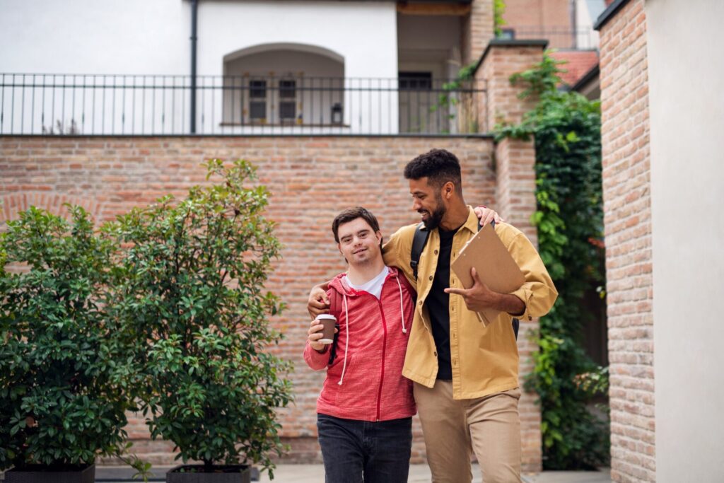 Young man with Down syndrome and his mentoring friend walking and embracing outdoors